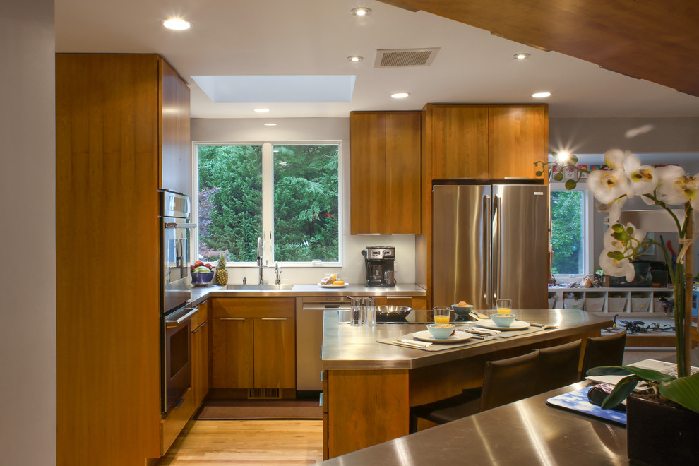 Small modern l-shaped eat-in kitchen in New York with an integrated sink, flat-panel cabinets, brown cabinets, stainless steel benchtops, white splashback, glass sheet splashback, stainless steel appliances, light hardwood floors, with island and brown floor.