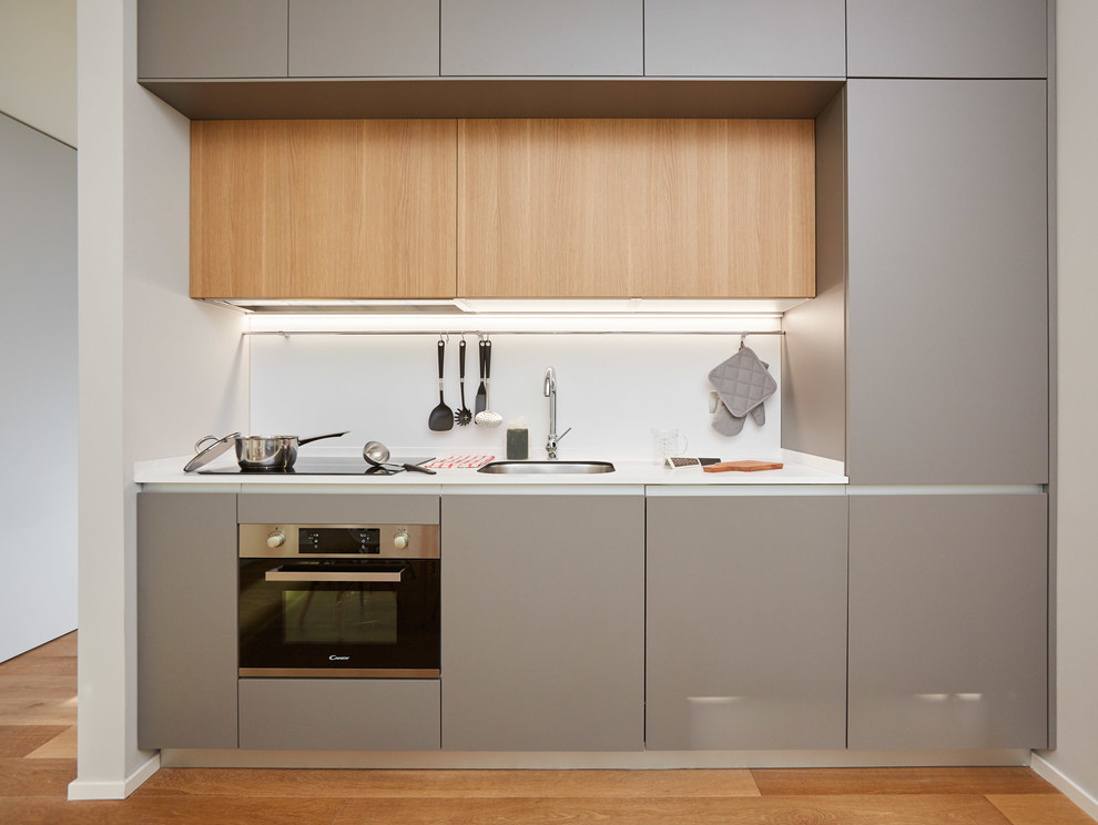 Small modern single-wall kitchen in Milan with an undermount sink, flat-panel cabinets, grey cabinets, white splashback, stainless steel appliances, medium hardwood floors, white benchtop, no island and brown floor.