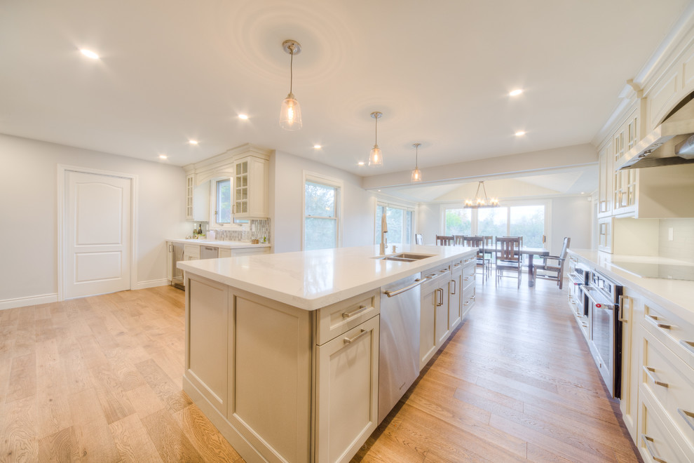 Example of a mid-sized transitional galley light wood floor and beige floor open concept kitchen design in Toronto with a double-bowl sink, recessed-panel cabinets, white cabinets, solid surface countertops, white backsplash, porcelain backsplash, stainless steel appliances, an island and white countertops