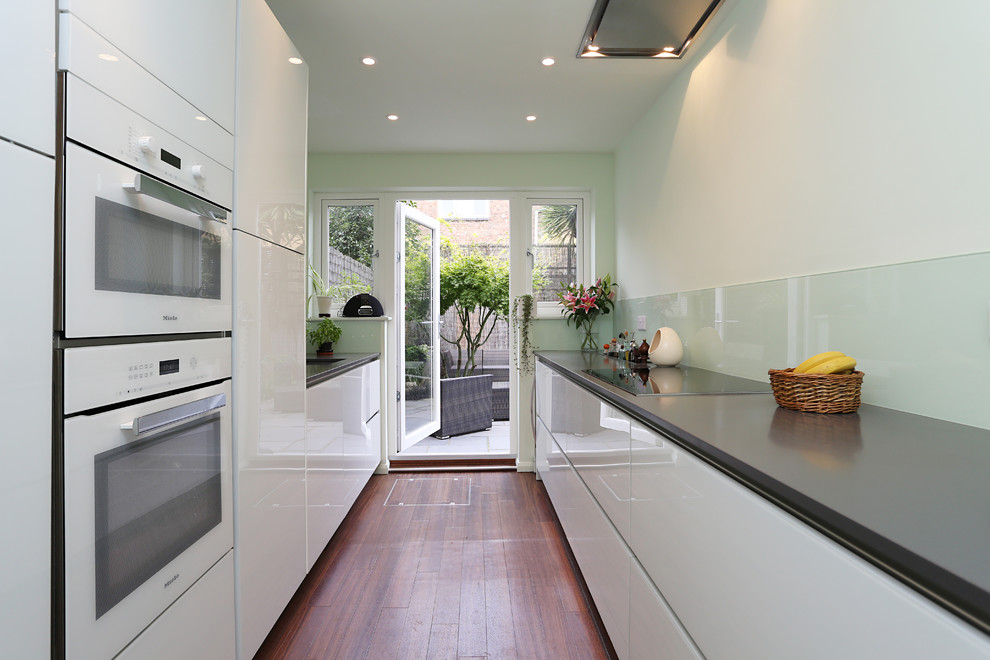 Small modern galley enclosed kitchen in London with a single-bowl sink, glass-front cabinets, white cabinets, quartz worktops, green splashback, glass sheet splashback, white appliances, dark hardwood flooring and no island.