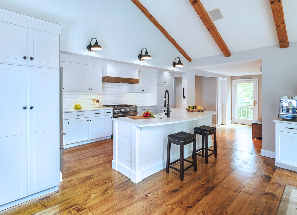 Mid-sized country galley open plan kitchen in Philadelphia with a farmhouse sink, recessed-panel cabinets, white cabinets, quartz benchtops, white splashback, subway tile splashback, stainless steel appliances, medium hardwood floors, with island, brown floor and white benchtop.