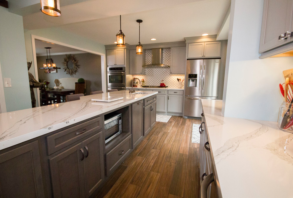 Inspiration for a large transitional l-shaped medium tone wood floor and brown floor enclosed kitchen remodel in Cincinnati with a farmhouse sink, recessed-panel cabinets, gray cabinets, marble countertops, white backsplash, porcelain backsplash, stainless steel appliances and an island