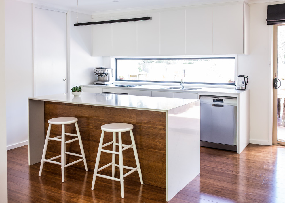 This is an example of a small modern galley open plan kitchen in Canberra - Queanbeyan with a double-bowl sink, flat-panel cabinets, white cabinets, quartz benchtops, window splashback, stainless steel appliances, bamboo floors, with island and brown floor.