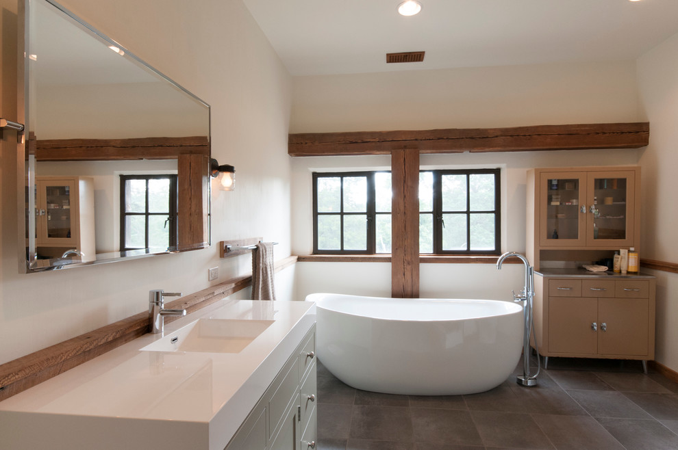 Photo of a country master bathroom in Other with furniture-like cabinets, light wood cabinets, a freestanding tub, stone tile, white walls, an integrated sink, porcelain floors, solid surface benchtops and brown floor.