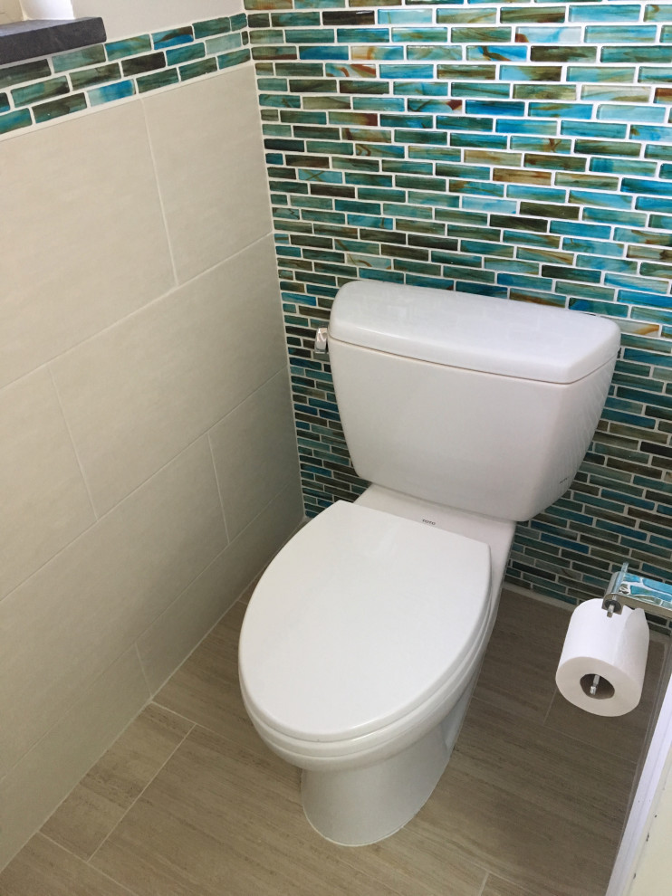 This is an example of a small beach style powder room in Orlando with glass-front cabinets, white cabinets, a two-piece toilet, blue tile, glass tile, black walls, porcelain floors, a pedestal sink, soapstone benchtops, beige floor and black benchtops.