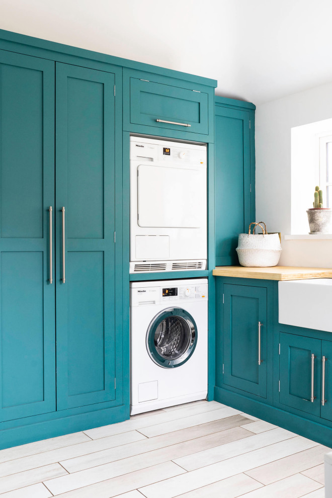 Design ideas for a small modern l-shaped dedicated laundry room in Hampshire with a farmhouse sink, shaker cabinets, turquoise cabinets, wood benchtops, white walls, vinyl floors, a stacked washer and dryer, white floor and brown benchtop.