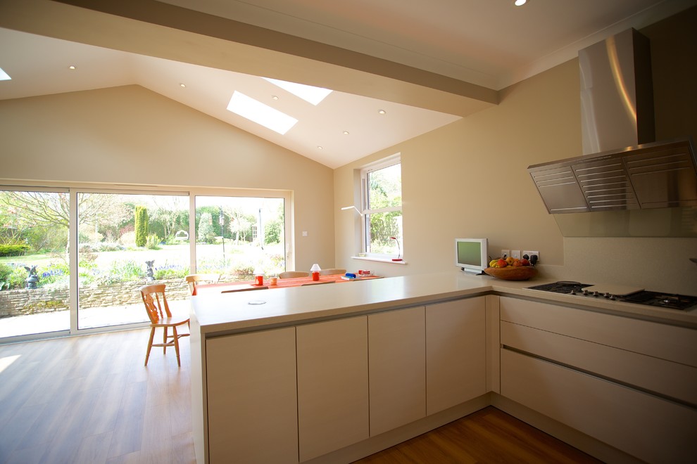This is an example of a mid-sized contemporary u-shaped eat-in kitchen in Hampshire with an integrated sink, flat-panel cabinets, white cabinets, solid surface benchtops, white splashback, stainless steel appliances, medium hardwood floors and a peninsula.