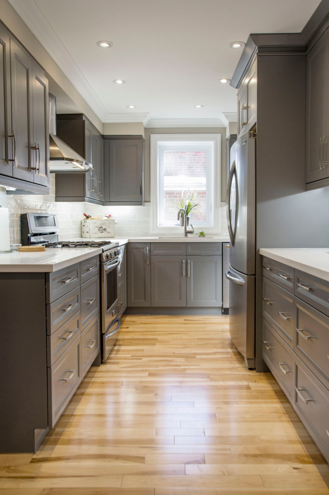 Example of a mid-sized transitional l-shaped medium tone wood floor and beige floor enclosed kitchen design with gray cabinets, quartz countertops, gray backsplash, ceramic backsplash, stainless steel appliances, white countertops, recessed-panel cabinets and no island