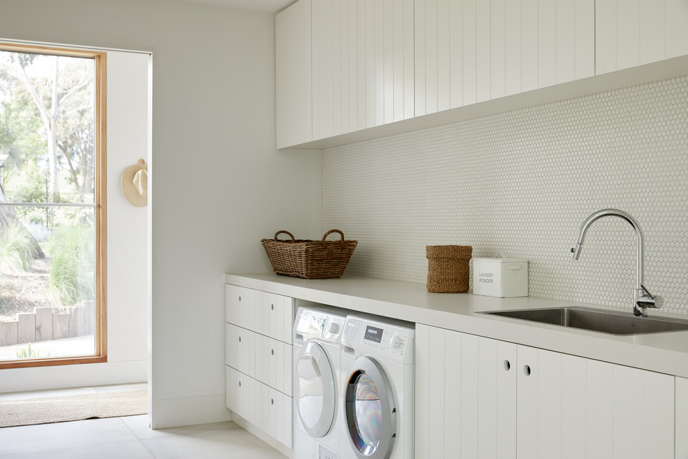 Photo of a scandinavian dedicated laundry room in Melbourne with a single-bowl sink, white cabinets, white walls, a side-by-side washer and dryer, grey floor and grey benchtop.