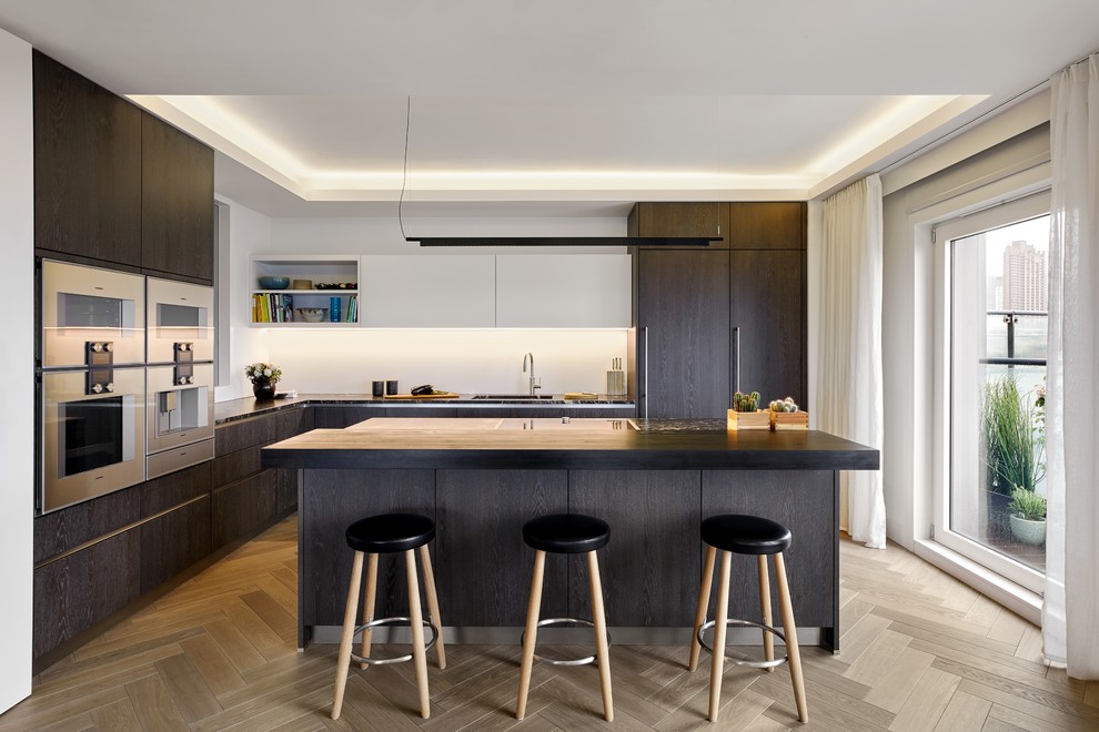 Design ideas for a mid-sized contemporary l-shaped kitchen in Gloucestershire with an undermount sink, flat-panel cabinets, dark wood cabinets, granite benchtops, white splashback, stainless steel appliances, light hardwood floors, with island, beige floor and black benchtop.