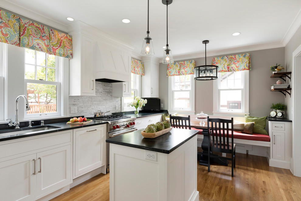 This is an example of a traditional eat-in kitchen in Minneapolis with an undermount sink, shaker cabinets, white cabinets, grey splashback, stainless steel appliances, light hardwood floors, with island and black benchtop.