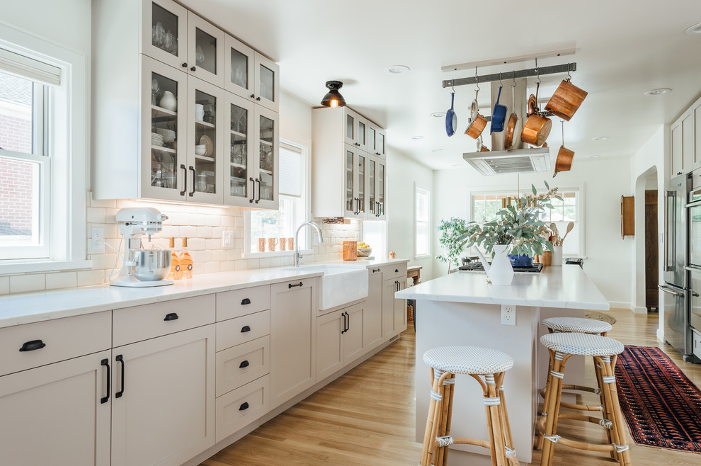This is an example of a large transitional galley eat-in kitchen in Salt Lake City with a farmhouse sink, glass-front cabinets, beige cabinets, solid surface benchtops, white splashback, ceramic splashback, stainless steel appliances, medium hardwood floors, with island and brown floor.