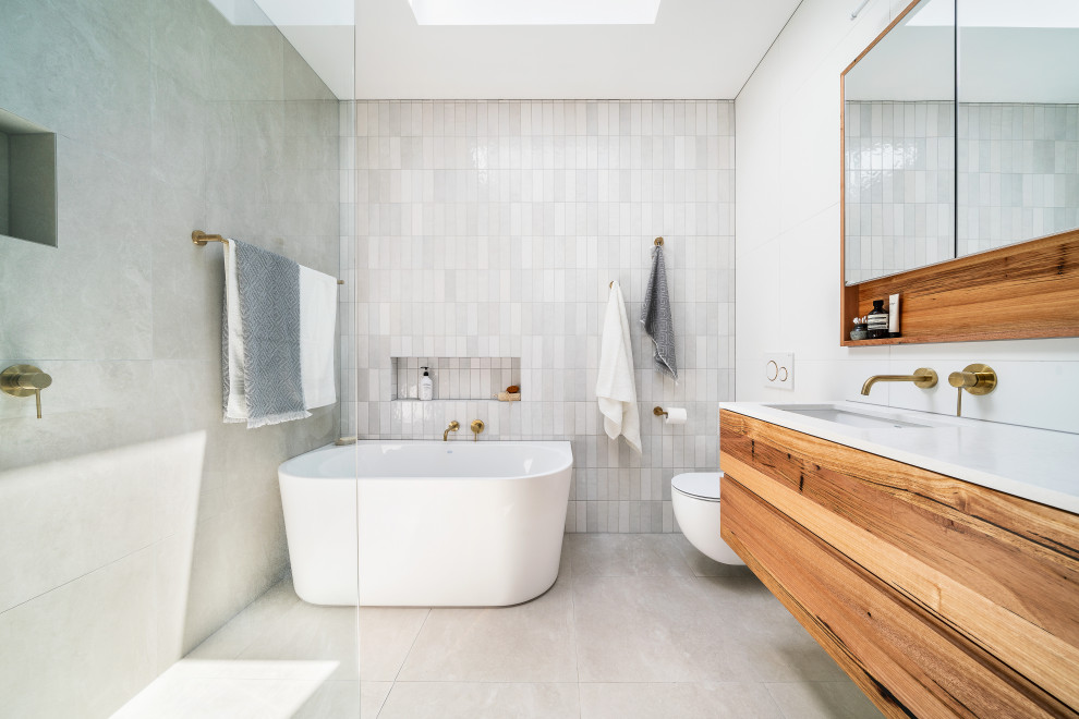 Bild på ett litet funkis vit vitt en-suite badrum, med skåp i mellenmörkt trä, ett fristående badkar, en öppen dusch, en vägghängd toalettstol, grå kakel, mosaik, vita väggar, klinkergolv i porslin, ett undermonterad handfat, bänkskiva i akrylsten, grått golv, med dusch som är öppen och släta luckor