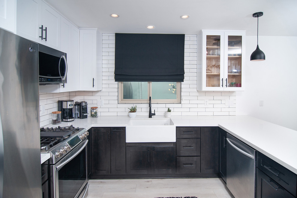 Photo of a mid-sized country u-shaped separate kitchen in San Diego with a farmhouse sink, shaker cabinets, black cabinets, quartz benchtops, white splashback, subway tile splashback, stainless steel appliances, light hardwood floors, a peninsula, brown floor and white benchtop.
