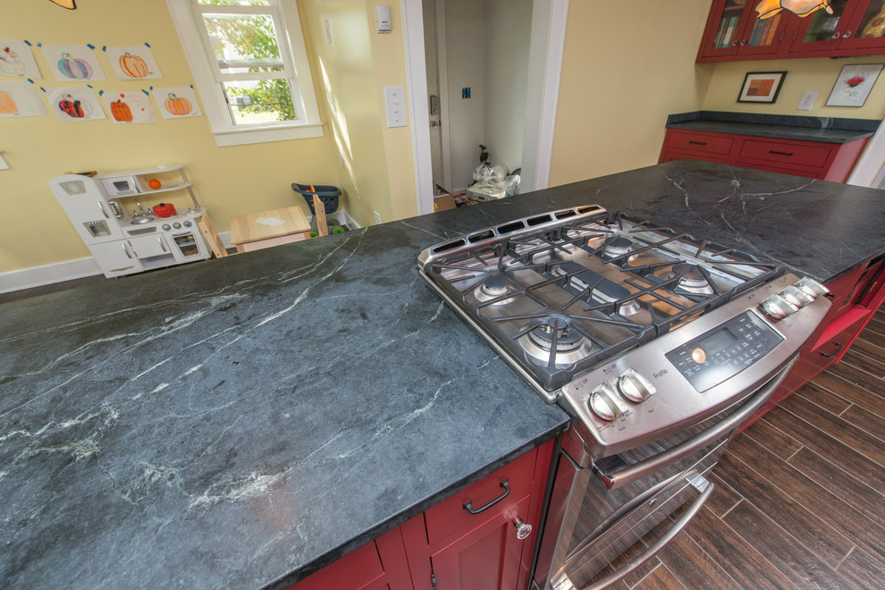 Mid-sized trendy galley dark wood floor and brown floor eat-in kitchen photo with a double-bowl sink, raised-panel cabinets, red cabinets, soapstone countertops, stainless steel appliances and an island