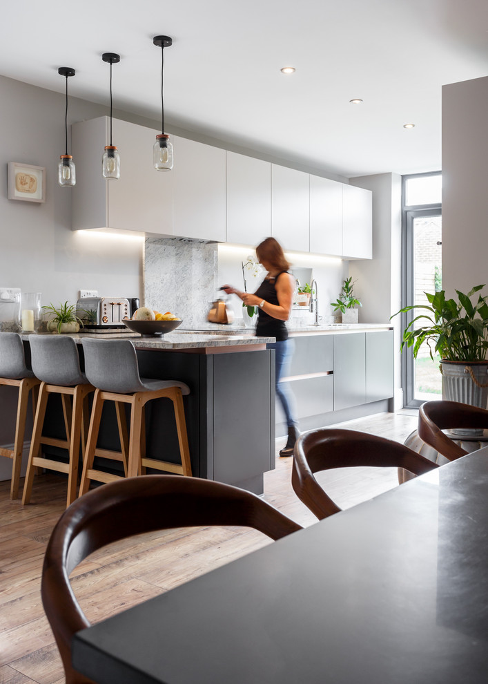 Inspiration for a contemporary l-shaped kitchen in Dorset with flat-panel cabinets, white cabinets, grey splashback, stone slab splashback, light hardwood floors, a peninsula and grey benchtop.