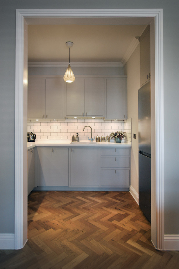 This is an example of a small modern u-shaped separate kitchen in London with an undermount sink, recessed-panel cabinets, grey cabinets, solid surface benchtops, white splashback, porcelain splashback, panelled appliances, medium hardwood floors, no island, brown floor and white benchtop.