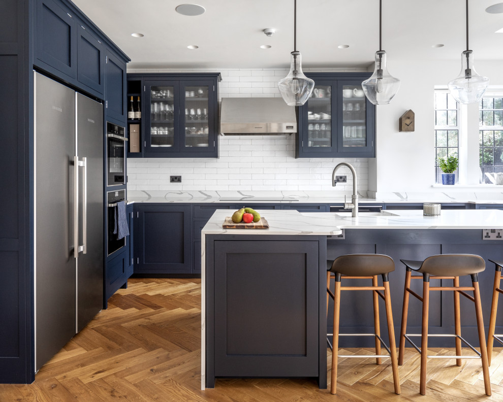 Design ideas for a mid-sized transitional l-shaped eat-in kitchen in London with a farmhouse sink, shaker cabinets, blue cabinets, quartzite benchtops, white splashback, porcelain splashback, stainless steel appliances, medium hardwood floors, with island, white benchtop and brown floor.