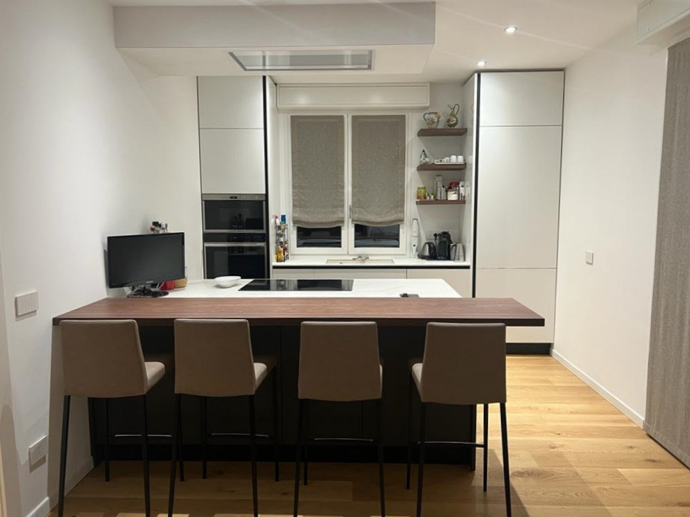 This is an example of a mid-sized contemporary galley open plan kitchen in Milan with an undermount sink, flat-panel cabinets, white cabinets, black appliances, light hardwood floors, a peninsula, white benchtop and recessed.