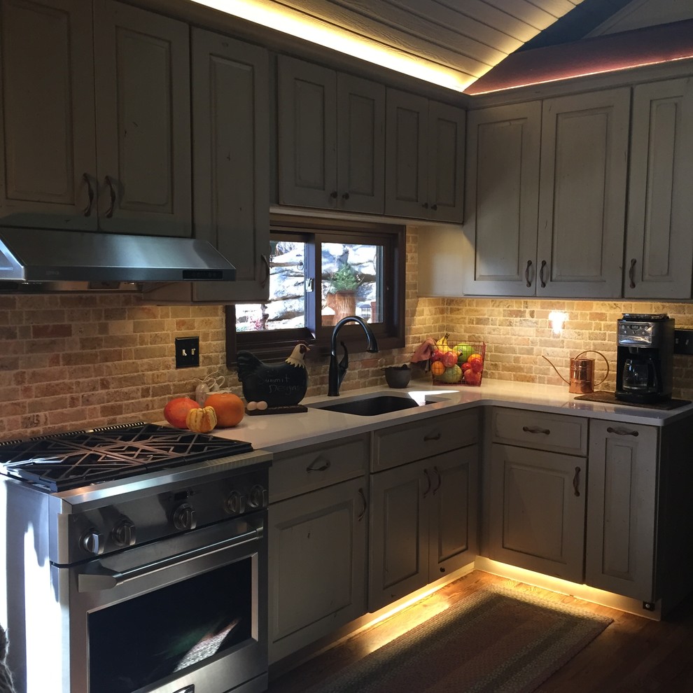 Example of a small mountain style l-shaped light wood floor kitchen pantry design in Denver with an undermount sink, raised-panel cabinets, distressed cabinets, stainless steel appliances, an island, solid surface countertops, stone tile backsplash and beige backsplash