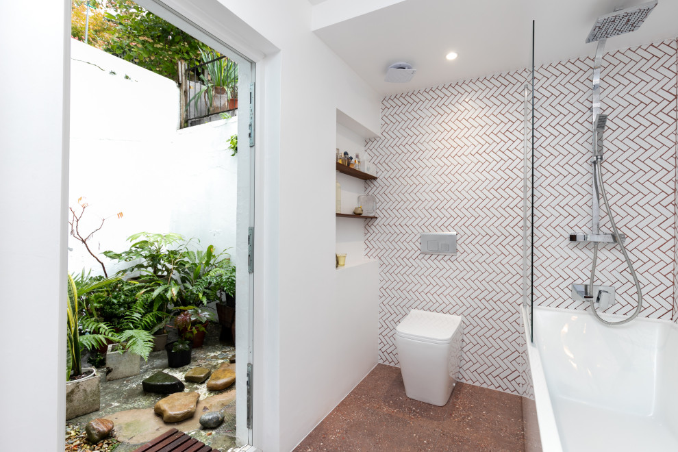 This is an example of a small modern kids bathroom in Sussex with brown cabinets, a freestanding tub, a shower/bathtub combo, a wall-mount toilet, white tile, ceramic tile, white walls, terrazzo floors, a wall-mount sink, solid surface benchtops, red floor, a hinged shower door, white benchtops, a single vanity, a built-in vanity, recessed and brick walls.