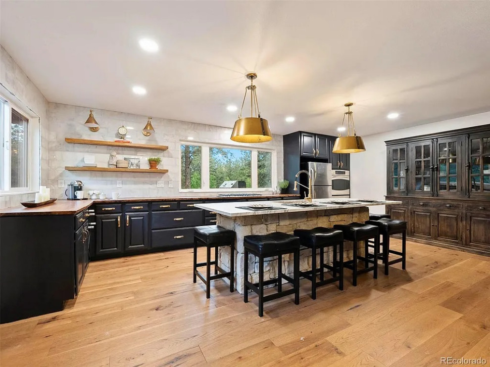 Open concept kitchen - large transitional u-shaped medium tone wood floor and brown floor open concept kitchen idea in Denver with an undermount sink, raised-panel cabinets, black cabinets, wood countertops, white backsplash, marble backsplash, stainless steel appliances, an island and brown countertops