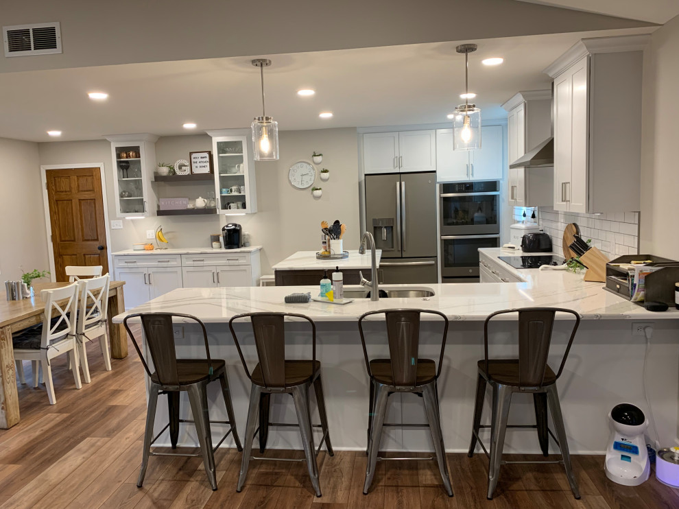 Open concept kitchen - large transitional u-shaped vinyl floor and brown floor open concept kitchen idea with an undermount sink, shaker cabinets, white cabinets, quartz countertops, white backsplash, ceramic backsplash, stainless steel appliances, an island and white countertops