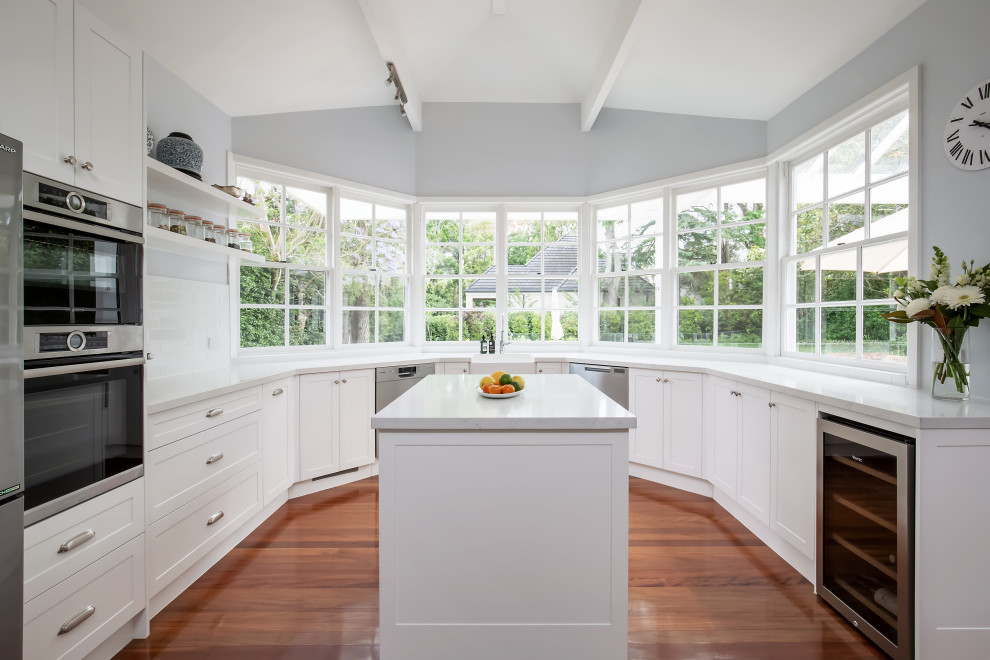 Inspiration for a large transitional u-shaped kitchen pantry in Sydney with a farmhouse sink, shaker cabinets, white cabinets, quartz benchtops, white splashback, subway tile splashback, stainless steel appliances, medium hardwood floors, multiple islands, brown floor, white benchtop and exposed beam.