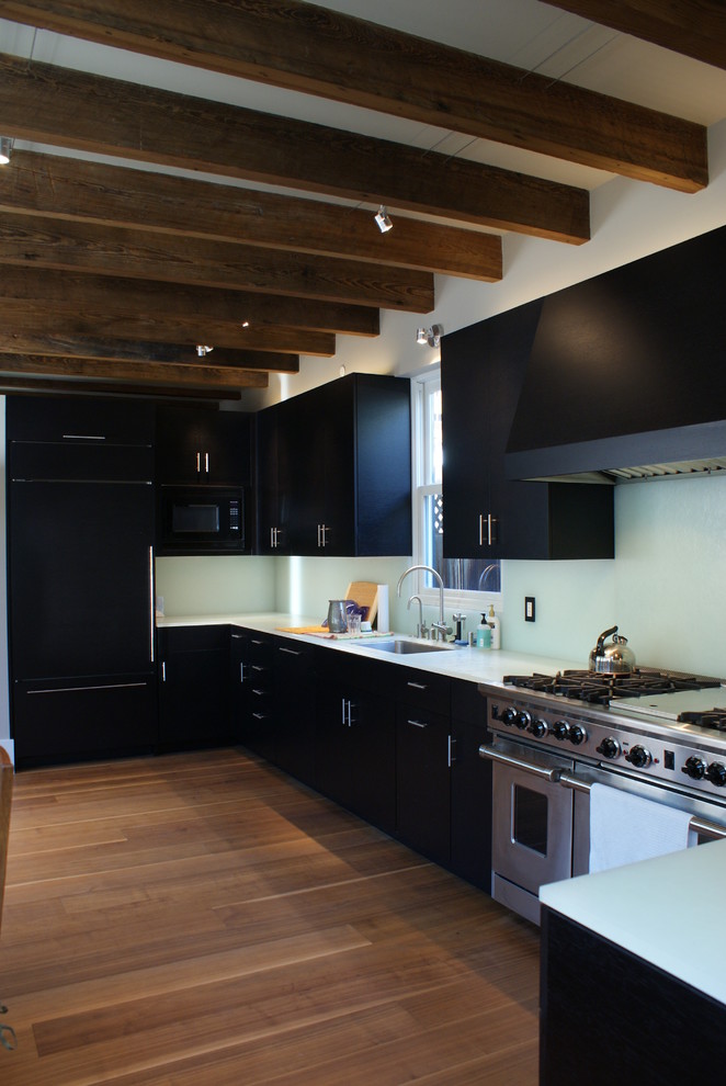 Photo of a mid-sized modern l-shaped eat-in kitchen in Denver with an undermount sink, flat-panel cabinets, black cabinets, glass benchtops, white splashback, glass sheet splashback, stainless steel appliances, dark hardwood floors and no island.