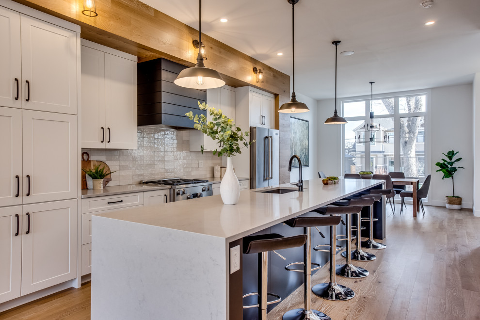 Example of a transitional galley medium tone wood floor and brown floor kitchen design in Calgary with an undermount sink, shaker cabinets, white cabinets, white backsplash, stainless steel appliances, an island and white countertops