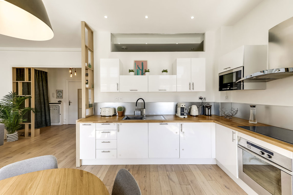 This is an example of a mid-sized scandinavian l-shaped eat-in kitchen in Paris with a drop-in sink, flat-panel cabinets, white cabinets, wood benchtops, stainless steel appliances, light hardwood floors, no island, metallic splashback, beige floor and beige benchtop.