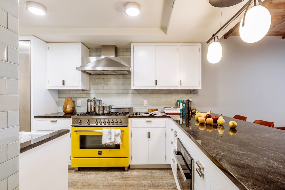 Photo of a large contemporary l-shaped open plan kitchen in New York with a farmhouse sink, flat-panel cabinets, white cabinets, quartz benchtops, white splashback, subway tile splashback, stainless steel appliances, light hardwood floors, a peninsula, brown floor, black benchtop and exposed beam.