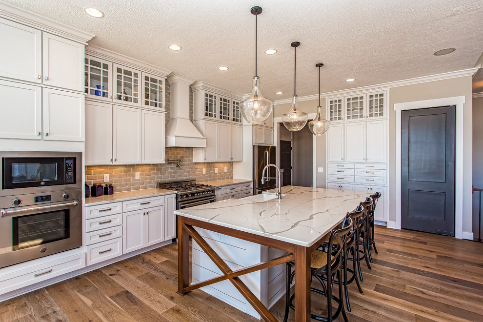 Inspiration for a traditional l-shaped kitchen in Cleveland with recessed-panel cabinets, white cabinets, grey splashback, subway tile splashback, stainless steel appliances, medium hardwood floors, with island, brown floor and white benchtop.