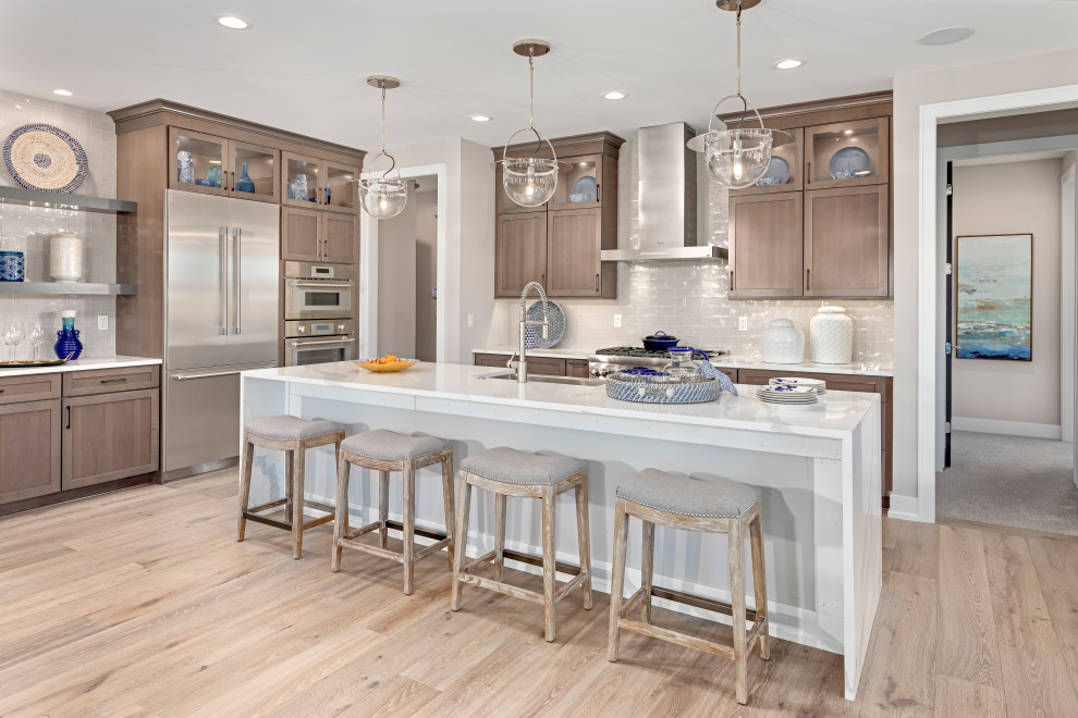 Photo of a transitional l-shaped kitchen in Detroit with a farmhouse sink, shaker cabinets, medium wood cabinets, grey splashback, stainless steel appliances, light hardwood floors, with island, beige floor and white benchtop.