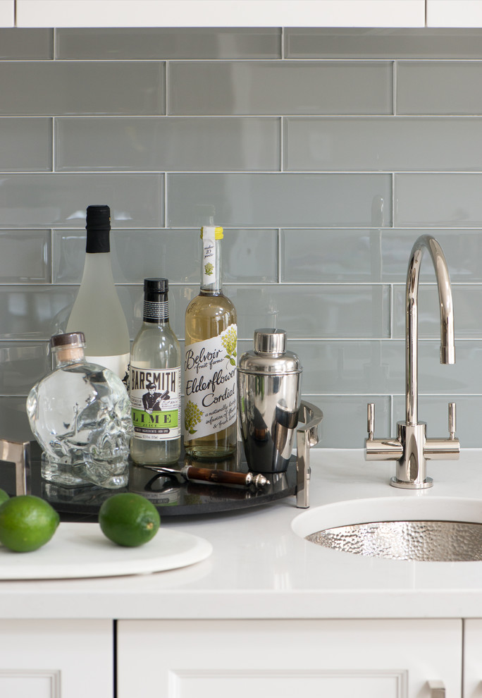 Photo of a small transitional single-wall wet bar in New York with an undermount sink, glass-front cabinets, white cabinets, quartz benchtops, grey splashback, glass tile splashback, porcelain floors, beige floor and white benchtop.