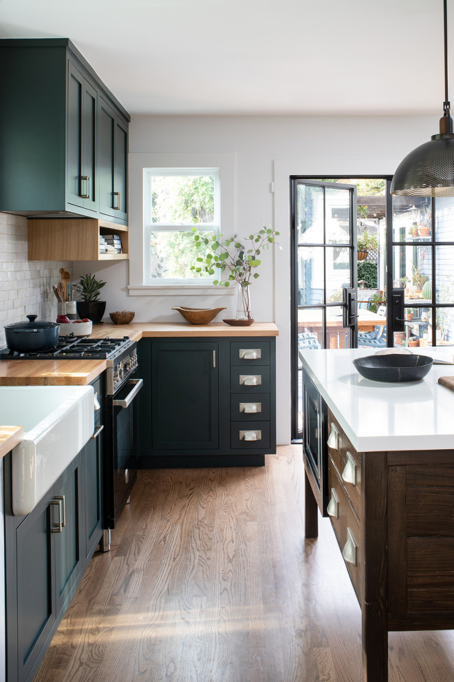 Photo of a mid-sized arts and crafts u-shaped separate kitchen in San Francisco with a farmhouse sink, shaker cabinets, green cabinets, wood benchtops, white splashback, terra-cotta splashback, black appliances, medium hardwood floors, with island, brown floor and brown benchtop.