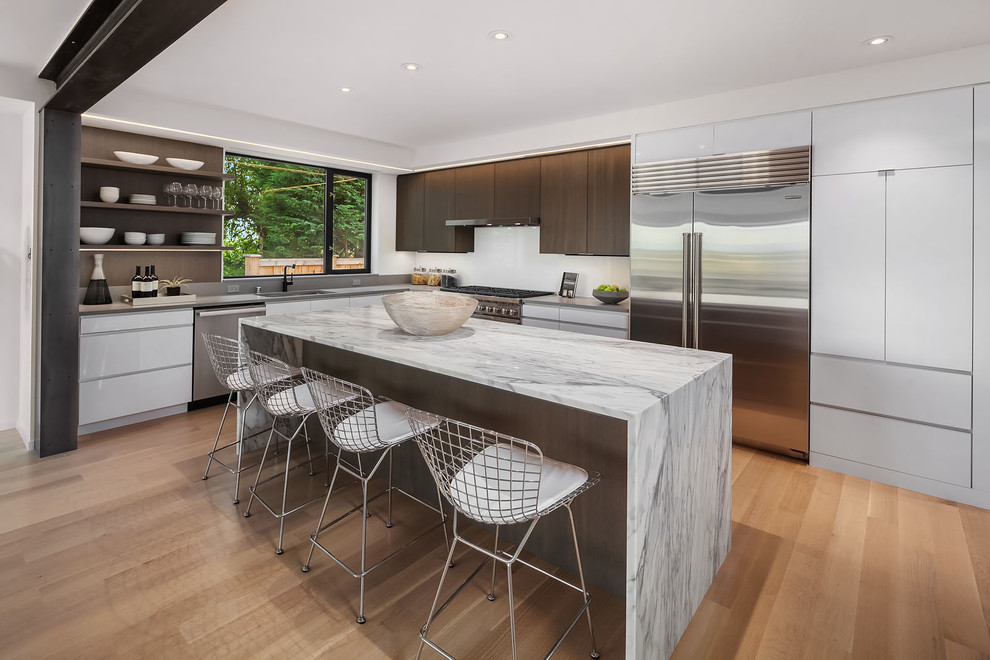 Photo of a mid-sized contemporary l-shaped open plan kitchen in Seattle with flat-panel cabinets, white cabinets, stainless steel appliances, light hardwood floors, with island, grey benchtop, an undermount sink, marble benchtops, grey splashback, stone slab splashback and brown floor.
