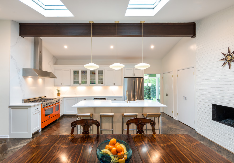 Photo of a mid-sized midcentury l-shaped eat-in kitchen in New York with a farmhouse sink, shaker cabinets, white cabinets, quartz benchtops, white splashback, stone slab splashback, coloured appliances, dark hardwood floors, with island, brown floor and white benchtop.