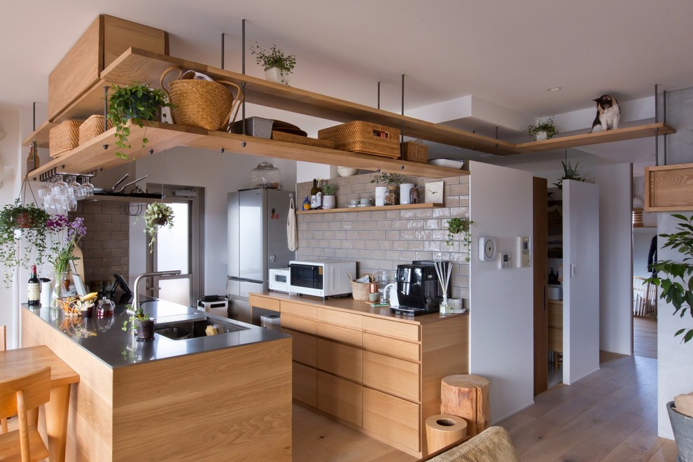 This is an example of a mid-sized country single-wall open plan kitchen in Other with an integrated sink, beaded inset cabinets, medium wood cabinets, stainless steel benchtops, white splashback, porcelain splashback, black appliances, medium hardwood floors, with island, beige floor and grey benchtop.