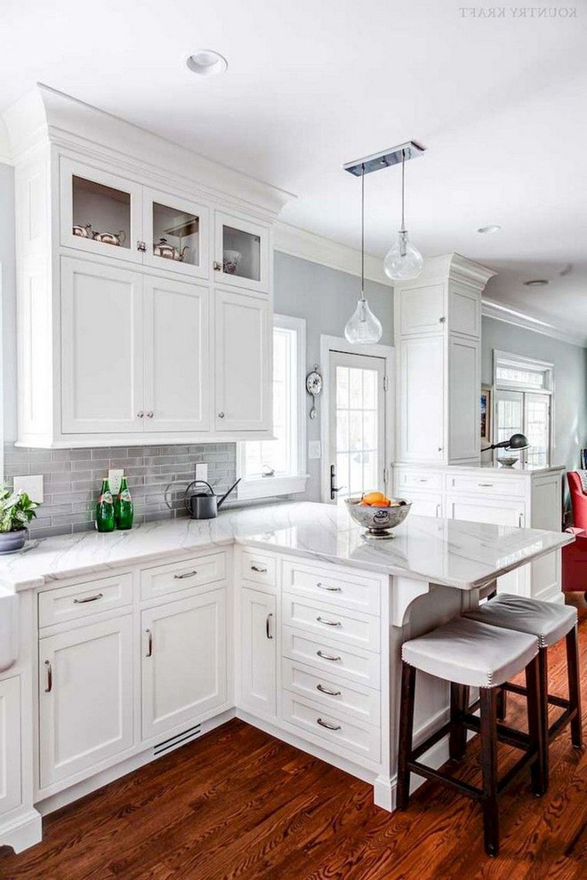 This is an example of a mid-sized traditional l-shaped eat-in kitchen in Columbus with a farmhouse sink, recessed-panel cabinets, white cabinets, marble benchtops, grey splashback, stone tile splashback, white appliances, dark hardwood floors, a peninsula, brown floor and grey benchtop.