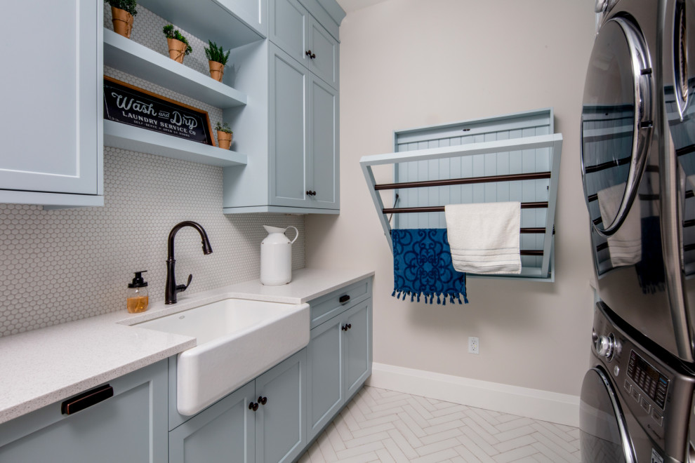 Photo of a mid-sized country galley dedicated laundry room in Toronto with a farmhouse sink, shaker cabinets, blue cabinets, quartz benchtops, grey splashback, ceramic splashback, beige walls, a stacked washer and dryer, white floor and white benchtop.