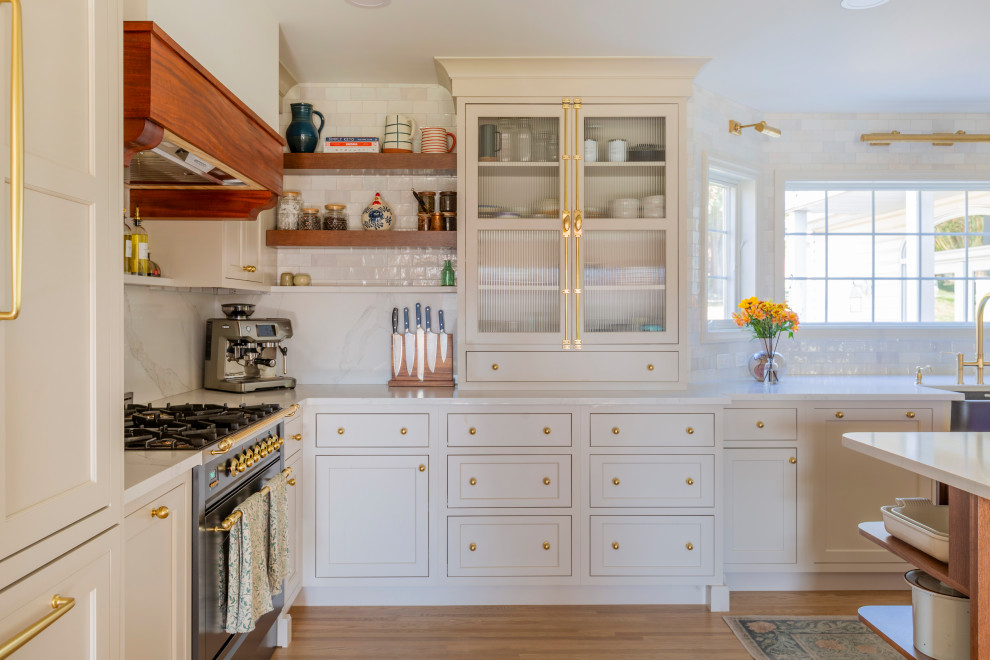 This is an example of a large traditional u-shaped kitchen pantry in New York with a belfast sink, shaker cabinets, white cabinets, engineered stone countertops, white splashback, ceramic splashback, integrated appliances, light hardwood flooring, an island and white worktops.