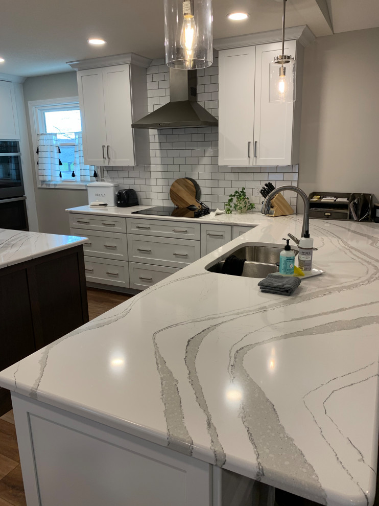 Example of a large transitional u-shaped vinyl floor and brown floor open concept kitchen design with an undermount sink, shaker cabinets, white cabinets, quartz countertops, white backsplash, ceramic backsplash, stainless steel appliances, an island and white countertops