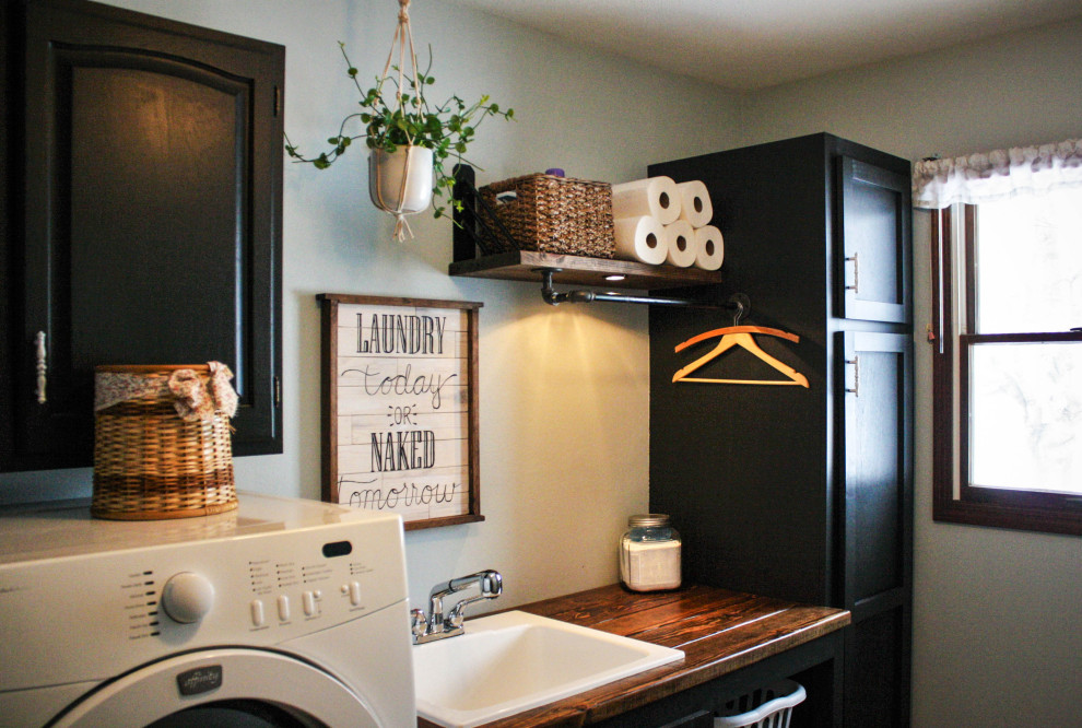 Photo of a mid-sized industrial single-wall dedicated laundry room in Other with an utility sink, shaker cabinets, black cabinets, wood benchtops, blue walls, linoleum floors, a side-by-side washer and dryer, beige floor and brown benchtop.