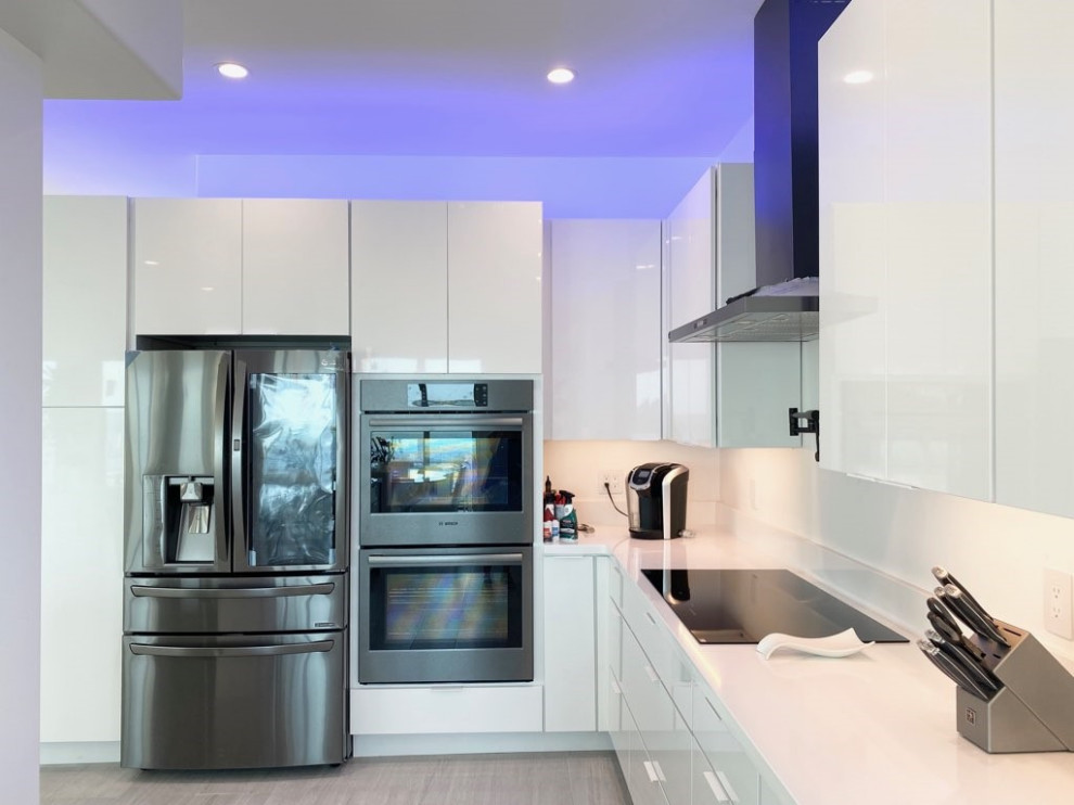 This is an example of an expansive contemporary l-shaped eat-in kitchen in Miami with an undermount sink, flat-panel cabinets, white cabinets, quartzite benchtops, white splashback, porcelain splashback, black appliances, with island, grey floor, grey benchtop and porcelain floors.