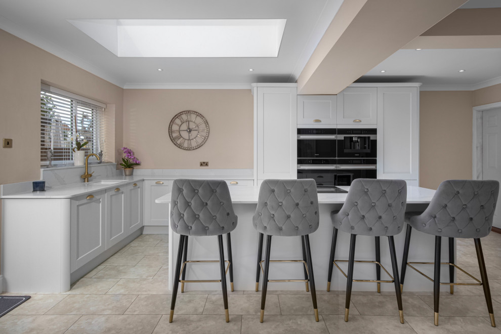 This is an example of a large country kitchen in Manchester with an integrated sink, shaker cabinets, white cabinets, quartzite benchtops, white splashback, black appliances, porcelain floors, with island, white benchtop and exposed beam.
