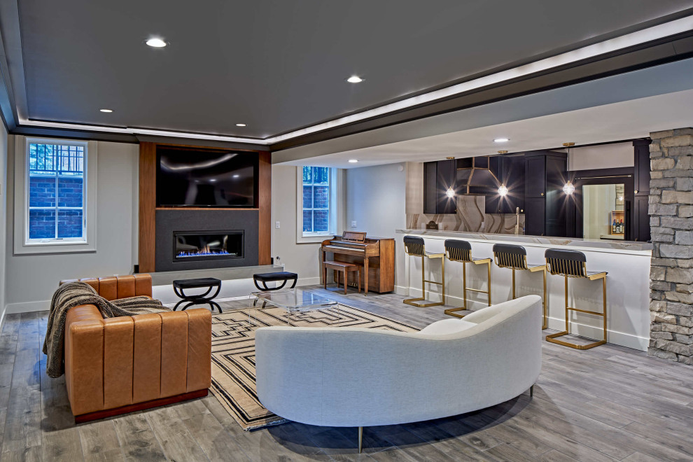 This is an example of a large contemporary fully buried basement in Detroit with a home bar, white walls, light hardwood floors, a standard fireplace, a wood fireplace surround and grey floor.