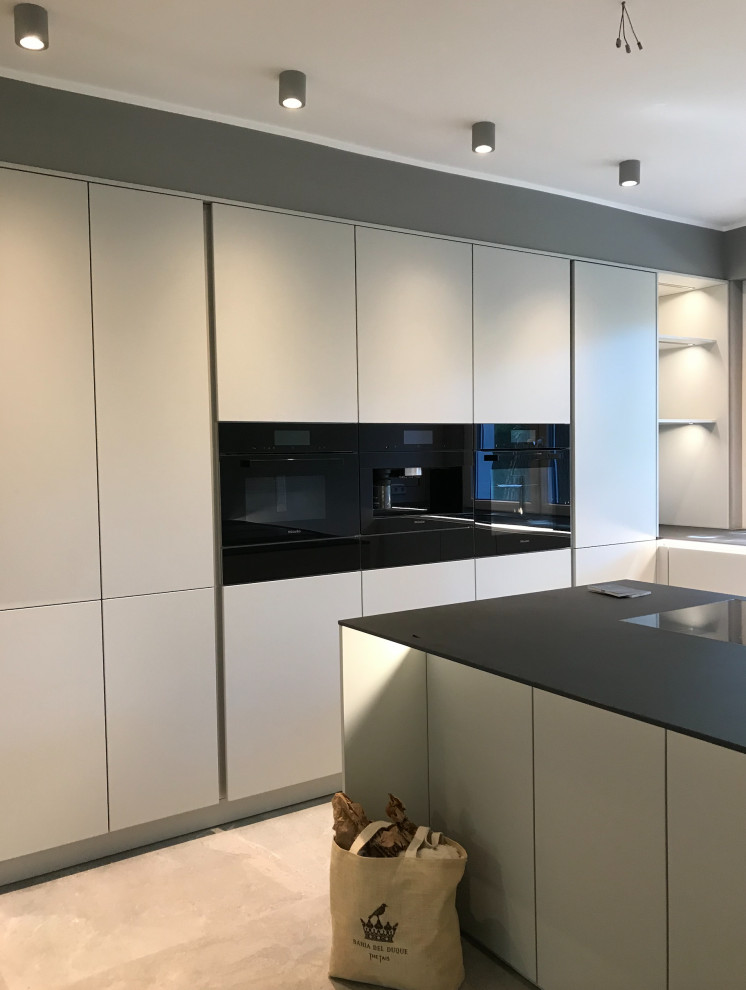 Large contemporary l-shaped open plan kitchen in Berlin with an undermount sink, flat-panel cabinets, black splashback, ceramic splashback, black appliances, ceramic floors, with island, grey floor and black benchtop.