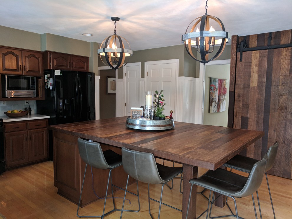 This is an example of a mid-sized country l-shaped kitchen in Other with raised-panel cabinets, medium wood cabinets, wood benchtops, grey splashback, subway tile splashback, black appliances, light hardwood floors, with island and brown floor.