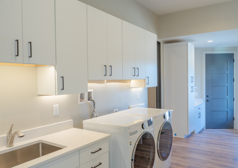 Photo of an expansive contemporary single-wall separated utility room in Denver with a submerged sink, flat-panel cabinets, white cabinets, quartz worktops, beige walls, light hardwood flooring, a side by side washer and dryer and white worktops.